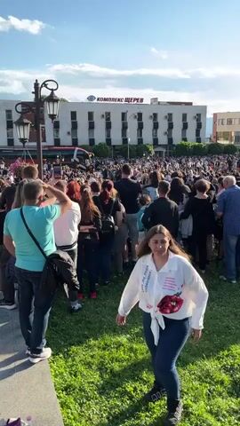 The video shows a large crowd of people gathered in an outdoor area, possibly a town square. The crowd is mostly facing towards the center of the area, with many holding up phones to record an event. In the background, there's a building with a sign in Cyrillic that reads "КОМПЛЕКС ШЕРЕВ", suggesting the location may be in Eastern Europe. A bus is parked near the front of the crowd, adorned with balloons, indicating a possible celebration or event. People are dressed casually in everyday attire. The weather is sunny with a few clouds in the sky. The environment includes grassy areas, trees, and street lamps, indicating a public space. The actions include people taking photos and videos, standing and watching, suggesting an organized gathering.