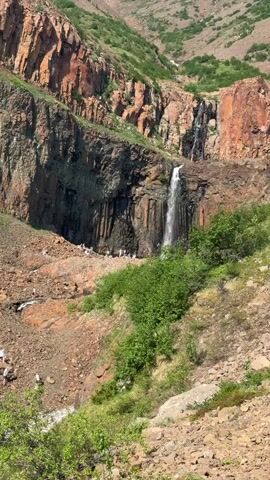 This video captures a scenic view of a rugged, mountainous landscape in Talnakh, Russia. The dominant features are sheer, reddish-brown rock cliffs, interspersed with dark, columnar rock formations that resemble organ pipes. A tall, slender waterfall cascades down the cliffs into a rocky ravine below.

Scattered across the rocky terrain, particularly in the middle ground and near the base of the waterfall, are several small groups of people. They appear to be dressed in light-colored clothing, likely for a hike or excursion in the area. Their presence suggests an activity such as sightseeing, hiking, or exploring the natural beauty of the location.

The scene is bathed in bright sunlight, indicating it is daytime, likely mid-morning or afternoon, given the shadows. The vegetation, predominantly green bushes and grasses, thrives on the slopes, suggesting a temperate climate. The overall impression is one of a remote and majestic natural environment, with human activity appearing small and incidental against the grand scale of the landscape. There is no visible text in the video.