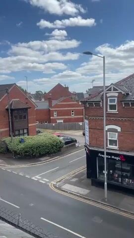 A view from an elevated position looking down at a suburban street. A two-lane road curves to the left, with two lanes of traffic. Several cars, including a white hatchback, a dark blue sedan, and a silver hatchback, are driving on the road. On the right side of the road, there is a row of brick buildings with tiled roofs. On the left side of the road, there are more brick buildings and some trees. The sky is blue with scattered white clouds. The weather appears to be sunny and fair.
