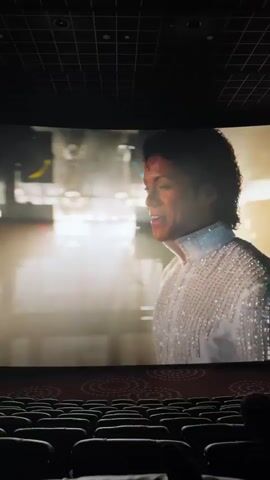 The video shows a movie screen in a theater displaying a scene with a person resembling Michael Jackson, wearing a sparkly white shirt. The person smiles and turns their back to the camera as they walk towards a brightly lit stage area. Subtitles in Chinese appear on the screen. The theater seats are visible in the foreground, mostly empty, with one person partially visible in the lower right corner. The ceiling of the theater is dark and has a grid pattern.