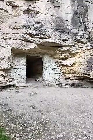 This video shows a path leading towards a cliff face. On the cliff face, there is an opening that appears to be a doorway. Further to the right, there is a stone structure built into the cliff with windows. A wooden cross stands in front of the stone structure. There are plants and trees growing around the area.