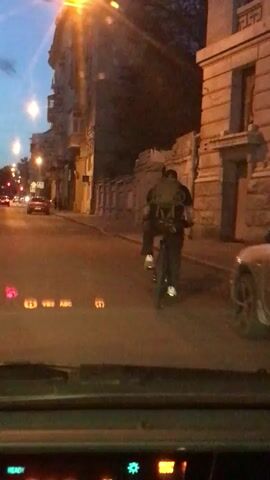 The video, taken from inside a car, shows a person riding a bicycle at dusk in a city street. The cyclist, seen from behind, wears a dark shirt, dark pants, and white shoes. They also wear a large backpack. The car is stationary, as the cyclist moves further away from the vehicle. The street is lined with buildings on either side. A few cars are parked along the street, and a red vehicle is visible in the distance. The road is paved with asphalt. The dashboard of the car is visible in the foreground, with illuminated displays showing various vehicle information.  The sky is a light blue, indicating the time of day.