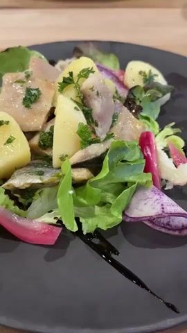 A close-up shot focuses on a dark gray plate filled with a salad. The salad contains chunks of cooked potatoes, pieces of fish, and green lettuce. Garnishes of pickled radishes, which are pink and purple, and chopped parsley are also visible. A dark drizzle, likely balsamic glaze, is artfully spread across the plate. In the background, out of focus, is another plate with french fries and a darker dish. The setting appears to be a restaurant with a wooden table.