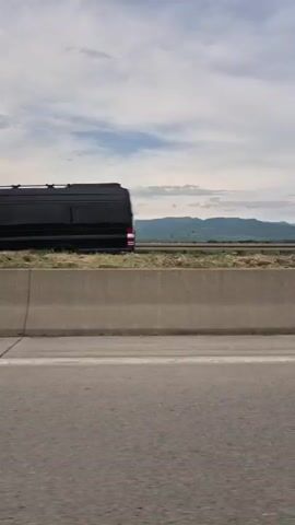 A car travels on a highway with a gray concrete barrier and a wide green median strip separating it from the other side. Low mountains are visible in the distance beneath a cloudy sky. As the car moves forward, a white van passes on the other side of the highway. The car's black side mirror is visible, reflecting the scene. The sound of wind is very loud.