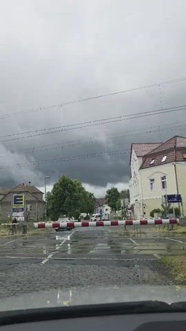 A video shows a closed railroad crossing in what appears to be a residential area. The crossing gate is down, indicating that a train is approaching or has passed. The road surface is wet, suggesting that it has recently rained. There are several buildings visible on either side of the road, including what appears to be a supermarket with an "E" logo. The sky is overcast and dark, with heavy clouds. There are power lines visible in the foreground. The video is shot from a vehicle.