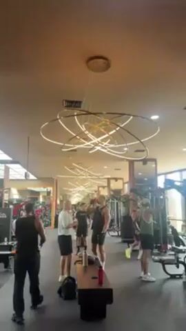 The video depicts the interior of a gym, with a focus on the ceiling. The ceiling features a series of modern, circular light fixtures, arranged in a layered, overlapping pattern. In the background, people are visible, some standing and talking while others appear to be working out. The gym equipment is visible in the background too. The overall atmosphere is that of a modern and well-lit gym environment.
