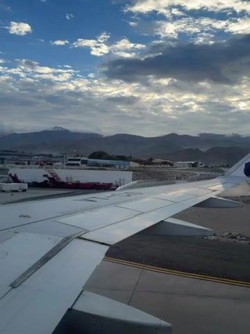 This video begins with a close-up view of an airplane wing, followed by a wider shot of an airport tarmac. In the background, several airplanes are parked, and a mountain range is visible under a cloudy sky. The camera then zooms in on a white airplane with red lettering and a staircase leading to its open door. Two crew members in red uniforms are standing at the top of the stairs. Luggage carts and other airport equipment are visible around the plane. The scene suggests boarding or disembarking is in progress. The video concludes with a shot of the airplane and the mountainous backdrop.