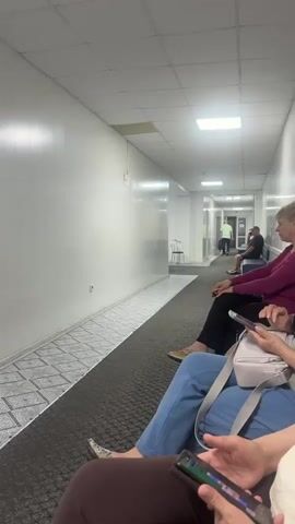 The video depicts the inside of a sterile-looking hallway, likely in a medical facility or similar institution. People are seated on benches that line one side of the hallway, most appearing to be waiting. At least three individuals are visible in the foreground, all looking at or interacting with their smartphones. The first person in the foreground is wearing blue pants, the second is wearing brown pants. The third is wearing a purple shirt and black pants. Further down the hall, a man in a green shirt is walking away from the camera, and another man is seated, wearing a hat and dark clothing. The hallway is brightly lit with overhead fluorescent lighting, and the walls are white and appear to be made of paneling. The floor consists of a dark gray carpet with a patterned tile border. A white plastic chair is visible at the end of the hallway.