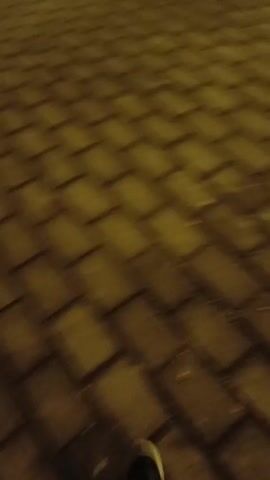 The video begins with a close-up shot of a brick paved street. The camera, unsteady and handheld, moves forward. The footage is lit with yellow light, and the camera moves upward to reveal the side of a building with an arched doorway and a tall window. Ornate decorations hang on the doorframe. An indistinct voice can be heard. The lighting is dim, making details difficult to discern.