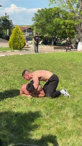 The scene captures two shirtless young men engaged in wrestling on a lush green lawn. One wrestler is on his knees, leaning over the other, who is lying face-down on the grass.  A scattering of onlookers observe from a distance, standing near trees and a row of benches. In the background are a house and a few parked cars, adding to the setting. Overhead, a blue sky with scattered clouds is visible.