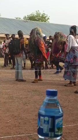 The video shows a person in a colorful, elaborate costume dancing in a dirt field. The costume consists of long, multicolored fringe covering their head and body, a patterned black top, and orange accents. A small crowd watches as the dancer moves, and other people in similar costumes can be seen in the background. A water bottle sits on the ground in the foreground. The event takes place outside, near a building with a corrugated metal roof.