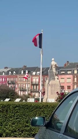 A French flag flutters on a flagpole in front of a statue of a soldier. In the background, buildings with red and pink facades line a street. Another flag, possibly Canadian, is visible on the left. A car's side mirror and part of its body are in the foreground on the right. A hedge runs horizontally across the bottom of the frame. The sky is clear and blue.