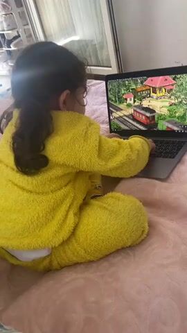 The video captures a young child, likely a toddler, seated on a bed. The child is dressed in a bright yellow, fuzzy hooded pajama set. The bed is covered with a soft pink blanket, and a blue and white striped pillow is partially visible to the child's left.

Initially, the child is seen from behind, engrossed in a cartoon playing on an open silver laptop placed on the bed. The laptop screen displays a colorful animated scene featuring a red train on tracks in front of a yellow house with a red roof, set in a green landscape. Bright sunlight streams in from a window or glass door in the background, indicating it is daytime.

As the video continues, the child changes position, bringing their legs up towards their chest. They then use both hands to bring their feet to their mouth, seemingly attempting to put them inside, all while occasionally looking back at the laptop screen. The child has dark, curly hair. The background visible through the window includes what appears to be shelves or storage units. The child appears both entertained by the cartoon and engaged in their own exploratory play.