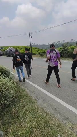 A group of four young men walk along a paved road with a white line marking the edge. They are walking away from the camera. The man in the foreground is wearing a purple and white t-shirt with the number 26 on the back and black pants. He is carrying a can in his right hand. The three men behind him are wearing black t-shirts and jeans. The man closest to the foreground is wearing a black t-shirt with white writing and blue jeans. He is holding a yellow bottle in his right hand. To his left, another man is wearing a black t-shirt with white writing and dark gray pants. The last man is wearing a black t-shirt and dark pants. In the background, there are several parked trucks and a vast expanse of lush green tea plantations stretching up a hill under a cloudy sky. The road is bordered by green grass and some tall, feathery plants on the left.