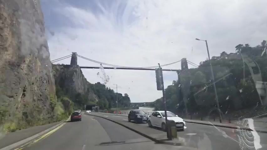 This media file captures a drive towards the iconic Clifton Suspension Bridge in Bristol, United Kingdom. The scene unfolds on a sunny day, with a partly cloudy sky suggesting fair weather. The perspective is from inside a vehicle, likely a car, as indicated by the dashboard and windshield reflections.

The primary subject is the Clifton Suspension Bridge, a magnificent feat of engineering spanning the Avon Gorge. Its grand suspension design, with two tall towers and thick cables, dominates the horizon. The surrounding landscape is dramatic, featuring steep, rocky cliffs on the left side of the road and lush green trees on the right. A river, likely the Avon, flows beneath the bridge, visible in the distance.

The road is a multi-lane carriageway, and several vehicles are in motion. A white car is in the foreground, to the right, its front end partially visible. Further ahead, a black car and a white car are driving in the same direction. On the left side of the road, a red car is also visible. The presence of road markings, streetlights, and traffic signals indicates an urban or semi-urban environment.

The time of day appears to be daytime, judging by the bright sunlight and clear sky. There are no people clearly visible in the frame, but the activity is clearly that of driving and commuting. The overall mood is one of observation and movement, as the viewer travels towards a well-known landmark. The media captures a dynamic view of the approaching bridge, emphasizing its scale and the natural beauty of its setting. No text is visible within the media itself.