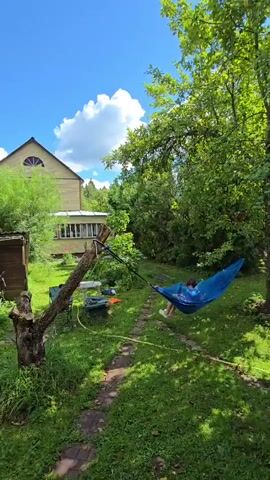 A person lies in a blue hammock suspended between a tree and a wooden post in a grassy backyard on a sunny day with a blue sky and white clouds. A two-story house with a screened porch is visible in the background. A path made of stepping stones winds through the grass. The hammock is positioned on the right side of the frame.