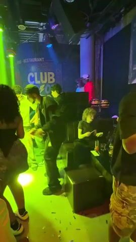 The video shows a group of people dancing in a crowded club. The lighting is colorful, with red, green, and blue lights shining across the room. There's a DJ playing music on a set of equipment in the background. One person is wearing a denim skirt and a black top. Another is wearing a green jacket and pants. The club appears to be a bar and restaurant, as indicated by a sign in the background. People are moving, dancing, and generally enjoying themselves in the lively atmosphere. Some are using their phones, while others are engaged in conversation or simply enjoying the music.