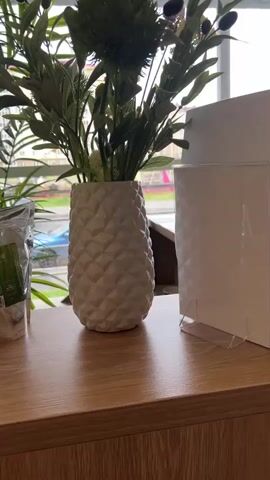 The video begins with a close-up of a white textured vase with greenery inside, placed on a wooden shelf. To the right, a white card is propped up in a clear stand. The camera then pans to the left, revealing a well-lit room with large windows. Inside, there are several potted plants arranged along a white radiator beneath the windows. A green welcome mat lies on the floor in front of a glass door. The camera continues to pan, showing a patterned rug on the floor and a gold shelving unit with various bottles and containers on its glass shelves. Further to the right, a black puffer jacket hangs on a gold coat rack. The camera then moves to focus on a distinctive, irregularly shaped mirror with an illuminated border, which reflects a woman standing in front of a reception desk in the background. Another potted plant is visible to the left of the mirror, and a white radiator cover with a leaf pattern is positioned below it. A dark green tufted bench is placed to the right of the coat rack. The woman in the reflection is holding a smartphone and appears to be taking a selfie. The overall environment suggests a modern, clean, and well-decorated interior space, possibly a salon or a boutique.