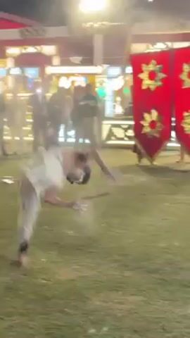 A man in a white dhoti and with black markings on his back is dancing barefoot on a lawn.  He is surrounded by people who are watching him.  Some are holding phones, filming him.  Behind the group of people, there is a building with a sign that says "Kapur Paar Palace."  To the right, a man in a black shirt and dark pants runs past two red banners with gold-colored designs.  He is holding a camera.