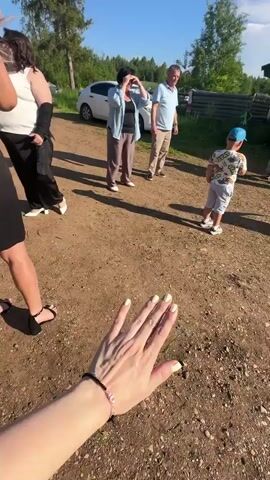 A group of people are gathered outdoors in a rural setting. In the foreground, a person's hand with manicured nails is visible, reaching towards the scene. A young boy wearing a blue cap and a patterned shirt stands in the middle of a dirt path, facing a group of adults. To the left, a woman in a black dress and heels is partially visible. Further back, several adults are standing in a semi-circle. Behind them is a small, dark wooden structure, possibly a gazebo or a small cabin, with a green roof. In the background, a white car is parked, and trees and a blue sky are visible. The adults appear to be interacting or observing the boy, with some clapping. The overall impression is a casual gathering or event taking place on a sunny day.