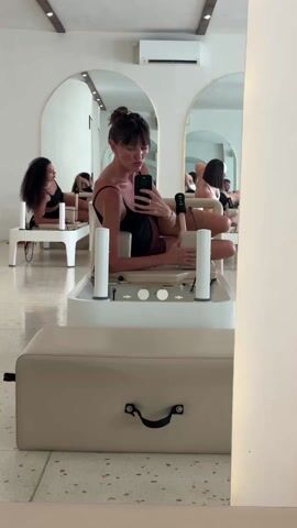 A woman is sitting on a pilates machine in a bright, white room. She is holding a smartphone and appears to be taking a selfie or video in the mirror in front of her. She is wearing a black top and maroon shorts, with her hair in a bun. There are other people on pilates machines in the background. The room has an air conditioner, arched mirrors, and light-colored speckled flooring.