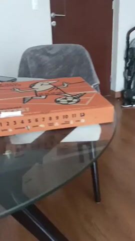 The video begins with a shot of a pizza box on a glass table. The box is orange and features cartoon imagery. A person's hand reaches into the frame and opens the pizza box, revealing a pepperoni pizza. The camera then zooms out slightly, showing more of the pizza and the interior of the box. The video ends with a shot of the pizza. The environment appears to be a room with wooden flooring and a door in the background. A stroller and shoes are also visible in the background.
