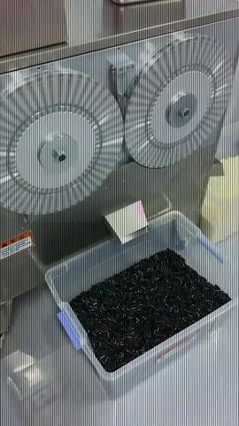 The video shows a clear plastic bin filled with small black objects, possibly pills or capsules, resting on a grey metallic surface. Behind the bin, a large, silver, industrial-looking machine is visible. This machine has two prominent circular components with radial grooves. There is a small white paper tag sticking out of the bin. The overall environment appears to be a clean, professional setting, likely a laboratory or manufacturing facility. The video is a close-up shot focused on the bin and the machine.