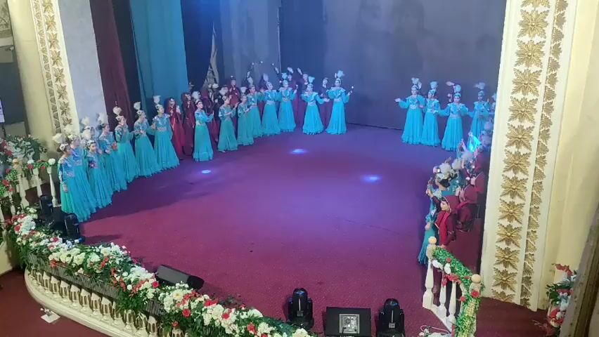 The video shows a group of young girls dressed in blue and red traditional outfits on a stage. They are performing a dance in front of an audience and a large screen displaying a group of people. The stage is decorated with flowers. The overall scene suggests a cultural event or performance.