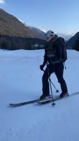 A person with ski gear, including a backpack, skis, ski poles, and goggles, is seen on a snow-covered ground, likely a ski area. The person is maneuvering on skis, using ski poles to move forward, against a background of snow-covered mountains, trees, and a clear blue sky. The person is wearing a cap, a jacket, and gloves. A white rope or fence can be seen in the background, marking boundaries in the snowy terrain.