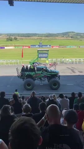 A monster truck with a green and black design is parked on an asphalt surface in front of a crowd of people seated in bleachers. The truck has four large wheels with green rims and a cage on the back with several people standing in it. A person in a green shirt is standing on the driver's side of the truck. Behind the truck is a grassy field with a race track and a building with stairs. The sky is blue and clear.