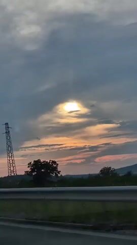 This is a vertical, medium shot video capturing a sunset sky viewed from a moving vehicle. The foreground features a section of a highway guardrail and the asphalt road. Beyond the guardrail, a grassy field stretches towards a distant treeline and rolling hills.

The dominant subject is the dramatic sky. Thick, gray clouds are interspersed with patches of lighter, wispy clouds. A bright, almost blinding sun is visible through a gap in the clouds, casting a golden glow that bleeds into soft oranges and pinks across the horizon. The light creates a halo effect around the sun.

In the mid-ground, a tall, lattice-style electricity pylon stands prominently against the sky, its dark silhouette stark. A single, broad-leaved tree is situated to the right of the pylon, with other vegetation forming a darker line along the hills.

The time of day is clearly sunset, evidenced by the warm colors in the sky. The weather appears to be partly cloudy, with the sun breaking through the overcast conditions. The location cues of rolling hills, vegetation, and the electricity pylon suggest a rural or semi-rural setting, consistent with the provided context of Peshterna, Bulgaria.

The overall mood evoked is one of expansive beauty and the transition from day to night, with a touch of industrial presence in the form of the pylon. There is no visible text in the video.