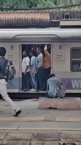 A train carriage with an open doorway is stationary at a platform. Five men stand inside the doorway, three facing left and two facing right. The men appear to be waiting to board or disembark. To the right of the train, a person is seated on a bench with a backpack. In the foreground, a person in a light-colored shirt and pants walks from left to right across the platform, also carrying a backpack. The platform is paved, and there are potted plants and station signage visible. The overall scene depicts a busy moment at a train station.