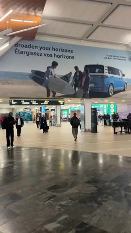 The media file captures an interior view of a busy airport terminal. The scene is illuminated by bright, overhead lighting, suggesting it's daytime. The overall atmosphere appears to be one of transit and movement.

In the foreground, the polished, dark tiled floor reflects the ambient light. In the mid-ground, several individuals are walking. Some are seen from behind, while others are walking towards the camera. Luggage, such as rolling suitcases, is visible, indicating people are traveling. A few figures are interacting near a grand piano, where a musician is playing, adding a touch of ambiance to the bustling environment. Beyond them, airport gates and signage are visible, with some people queuing or walking through doorways.

A prominent advertisement stretches across the upper portion of the scene. It features a blue Volkswagen ID. Buzz electric vehicle parked on a beach with surfboards. The advertisement's text reads, "Broaden your horizons. Élargissez vos horizons." Below this, smaller text indicates it's an "All-Electric ID. Buzz Ltd. Buzz 100% Électrique." This advertisement provides a visual clue about the airport's potential location, possibly an area where VW's advertising is prevalent or a location where outdoor activities like surfing are popular.

The presence of airport signage, the movement of travelers with luggage, and the overall layout of the space confirm the setting as an airport. The time of day is suggested to be daytime due to the bright interior lighting. No specific weather conditions are discernible from the indoor view. The general mood appears neutral, consistent with a functional transportation hub. The media file appears to be taken in Richmond, Canada.