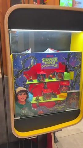 The video shows the interior of a McDonald's restaurant. Initially, the camera focuses on a display case containing "Stranger Things" Happy Meal toys. The display case is colorful and features images of the show's characters and the "Upside Down" world. Then, the camera pans to the right, revealing the McCafe section of the restaurant. There's a display case filled with pastries and desserts, and a barista is visible behind the counter, preparing drinks. The interior design is modern, with a combination of wood and white surfaces. The restaurant appears to be clean and well-lit.
