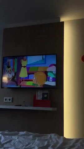 The video captures an indoor scene, likely a modern hotel room or bedroom, at night or in dim lighting. A flat-screen television, mounted on a wooden-paneled wall, is playing an animated show featuring characters in what appears to be a clothing store setting. A "30%" discount sign is visible on the TV screen during one animated frame. Below the TV, a white shelf is affixed to the wall, holding two electrical outlets on the left and a red box with some papers inside on the right. A white bed with a textured, striped duvet occupies the lower foreground of the shot. To the right of the TV, a vertical strip of warm yellow light illuminates a beige wall, and a small, red circular object is mounted on the wall further right. A bedside lamp with a warm glow is partially visible on the left side, beside the television. The room's artificial lighting creates a cozy, relaxed atmosphere.