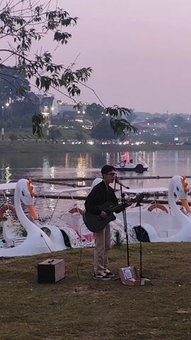 A young man with glasses is playing an acoustic guitar and singing into a microphone on a grassy area next to a lake at dusk. He is dressed in a dark jacket and light pants. Behind him are several swan-shaped pedal boats docked on the lake, their white forms contrasting with the darkening water. In the distance, city lights twinkle, reflecting on the water's surface. Another person walks behind the swan boats on the right side of the frame. The scene is calm and peaceful, with the focus on the musician's performance in a scenic setting.