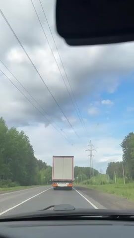The video captures a forward-facing view from inside a moving vehicle, likely a car, driving on a two-lane asphalt road. In the foreground, the top of the dashboard and the housing of the rearview mirror are visible. The primary subject is a large white and red semi-trailer truck, which the camera vehicle is following.

The road is flanked by dense green trees on both sides, suggesting a rural or semi-rural setting. Overhead, numerous power lines stretch across the sky, converging towards a tall metal power pylon visible on the right side of the road in the distance. The sky is partly cloudy, with significant grey clouds dominating the left side of the frame and clearer blue sky with lighter clouds towards the right.

The truck's rear doors are white, and its lower bumper area is red. A license plate is partially visible on the truck's rear, but the characters are not decipherable. Road markings, including solid white lines and dashed white lines, are present on the asphalt. Two small circular road signs are visible on the right side of the road, partially obscured by foliage and distance, making their specific messages unreadable. The overall scene depicts a daytime journey through a wooded area under a mixed sky. The location context indicates this is in Pavlovskii, Russia.