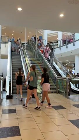 The video shows an indoor scene, likely a shopping mall, with people using escalators and stairs. In the foreground, four young females are walking up a set of stairs. One is wearing a green t-shirt and black shorts, another a black t-shirt and pink shorts, and the third a black t-shirt and black shorts. The fourth person is mostly out of frame but appears to be wearing dark clothing. Behind them, the escalators are visible, with several people ascending and descending. On the escalator to the left, a man and woman are ascending, followed by a woman with a large bag. On the escalator to the right, a man in a grey shirt and black pants is ascending, while further up, a group of people are descending, including a woman in a pink and white floral dress. The stairs themselves are decorated with a colorful, abstract pattern. The environment appears bright and well-lit, with polished floors and glass railings.