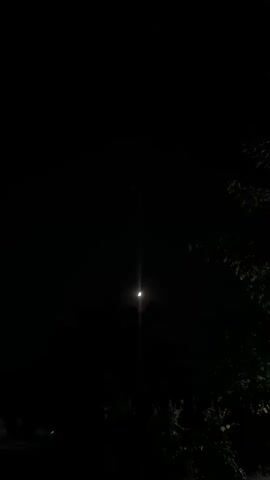 A video captures a crescent moon in a dark night sky. The moon is illuminated from the right, showing a bright sliver of light against the blackness. A bright, vertical light beam emanates from the moon, creating a lens flare effect. Silhouetted trees or foliage are visible at the bottom of the frame, obscuring the ground. The overall scene is dark, with the moon being the primary light source.