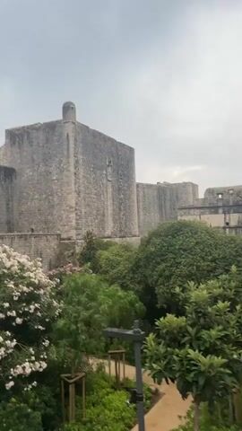 This media file captures a view of the historic city walls of Dubrovnik, Croatia, on a cloudy day. The imposing stone fortifications, characteristic of Dubrovnik's Old Town, dominate the upper portion of the frame. Lush green trees and bushes, including a flowering bush with white and pink blossoms, are visible in the foreground and mid-ground, adding a vibrant contrast to the ancient stone. The scene appears to be during daylight hours, given the overall illumination, though the overcast sky suggests it might be early morning or late afternoon, or simply a day with diffused light.

The camera perspective is from a walkway, possibly along the city walls themselves, overlooking a path where a few people are walking. One individual is seen from behind, wearing a dark top and light-colored pants, carrying a folded umbrella. Another person, wearing a blue top and shorts, is walking ahead. These individuals suggest that the location is accessible to tourists or residents. A small flag, appearing to be the Croatian flag, flies from a tower atop the city walls, providing a clear location cue.

The overall mood conveyed is one of historical grandeur and the peaceful coexistence of nature with ancient architecture. The texture of the stone walls is palpable, hinting at centuries of history. The presence of greenery and the path suggest a well-maintained tourist attraction. No visible text is present in the media file.
