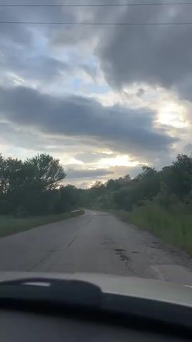 The video shows a first-person perspective from inside a vehicle driving on a cracked, asphalt road. The road winds uphill through a rural area with trees and vegetation on either side. The sky is overcast with dark, heavy clouds, but a patch of brighter sky and sunlight peeks through the horizon. The vehicle's dashboard and windshield are visible at the bottom of the frame.