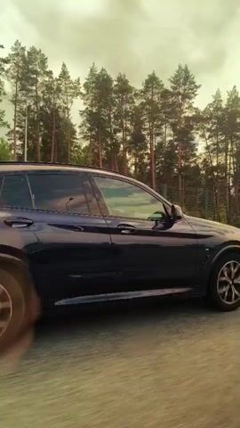 A dark-colored BMW X4 SUV drives along a paved road. The vehicle is seen from the side and rear, with a backdrop of a dense forest and a cloudy sky. The road appears to be a highway with a grassy median. The car's license plate is visible as "JU 5815". The video is shot from a low angle, giving a dynamic perspective of the car in motion.