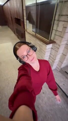 A young woman with red hair, wearing glasses and black Marshall over-ear headphones, records a selfie video while walking through an indoor corridor. She is dressed in a red long-sleeved top and dark red or maroon pants. The woman is smiling and appears happy and playful, at one point extending her arm.

The setting is a hallway with light grey tiled floors. On one side, there's a wall with a brick-like pattern, featuring a window or glass partition and a sign with the letter "B" visible. The opposite side of the hallway has wooden paneling and doors. The architecture suggests an older building, consistent with the provided location context of Sanremo, Italy.