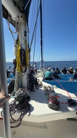 The video shows a sailboat moving across the ocean under a clear blue sky. A group of people is sitting on a blue net, enjoying the ride. There's a mast with ropes and rigging.  On the deck are shoes and gear. Winches are visible, with ropes coiled around them.