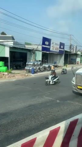 The video shows a busy street scene in Vietnam. A large white semi-truck with a blue cargo container is seen driving along the road. Several motorcycles are seen moving along the road and crossing the intersection. There is a building with a sign in Vietnamese in the background. The road surface is paved but appears worn in places. The sky is clear.
