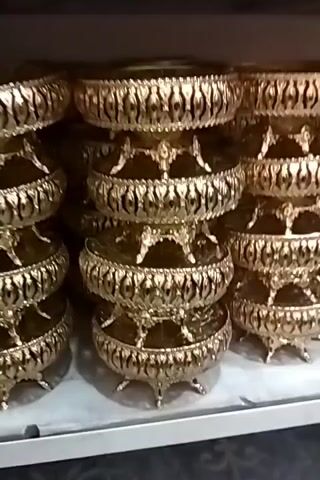 The video shows a display of ornate, gold-colored decorative bowls stacked on shelves. The bowls appear to be made of metal and have intricate designs on their rims and stands. They are arranged in multiple columns and rows, suggesting they are for sale or display in a store. The lighting is somewhat dim, and the background is out of focus, further emphasizing the product. There are no people or animals visible in the video.