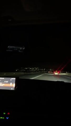 The video shows a view from inside a car at night, looking out the windshield. The car is on a highway, with lights from a distant city visible on the horizon. Other cars with their headlights on are also visible on the road ahead. The dashboard of the car is dimly lit, with a digital display showing what appears to be audio information. There is also a red laser-like light emanating from the right side of the windshield. The road is dark and the sky is black.