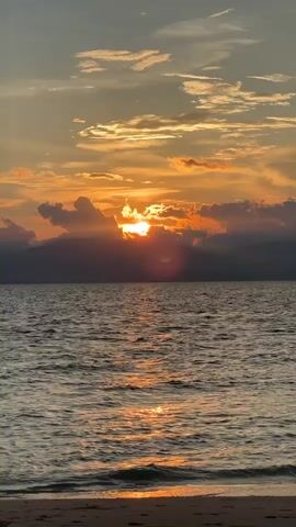 The video shows a sunset over a body of water. The sun is partially obscured by clouds, casting orange and yellow light across the sky. The water is choppy, with waves breaking on a sandy shore in the foreground. Mountains are visible in the distance.