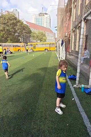 A young child wearing a blue and yellow soccer uniform stands on a green turf field. The child is facing forward and appears to be looking down. In the background, two yellow school buses are parked. Other children in similar uniforms are visible near the buses. The environment appears to be an outdoor sports field with buildings and trees in the distance. A soccer net is visible to the right of the child. The sun is shining, casting shadows on the field.