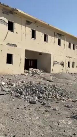 The video shows a severely damaged two-story building and a destroyed vehicle, likely a station wagon. The building has multiple bullet holes and significant structural damage, including missing sections of the walls. Windows are broken and absent. Debris is scattered around the base of the building. In front of the building, there is a heavily damaged white car, with its windows broken, hood crushed, and body riddled with holes. The surrounding area is covered in rubble and debris, indicating a scene of conflict or destruction. There's no sign of people in the frame. The sky is clear.